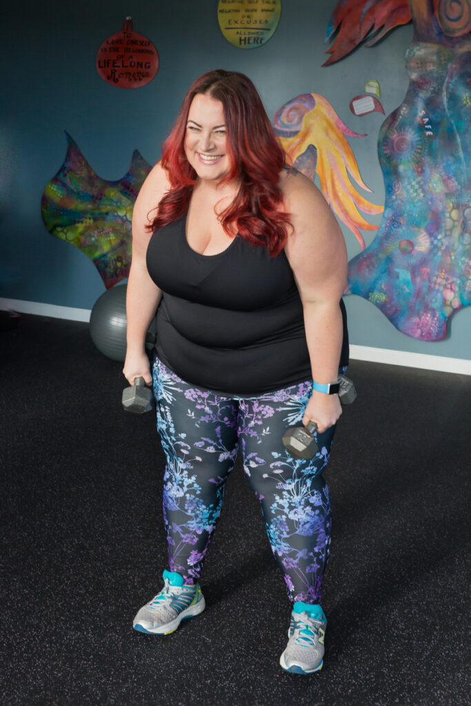 A red-haired, plus size woman poses with hand weights in soft light in a fitness studio converted from a garage. This woman is a professional body positive fitness instructor who practices health at every size (HAES). She's wearing a black tank top and colorful blue and purple leggings, and is looking directly at the camera and smiling.